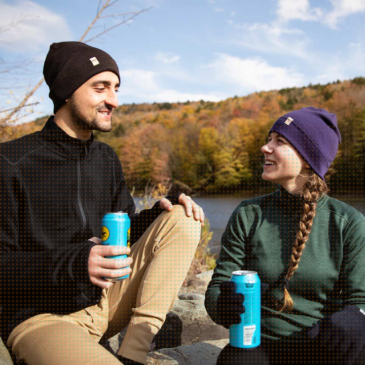 Two people sit on a rock engrossed in conversation in front of a mountain-lined lake. The man models the lightweight beanie, heavyweight full zip sweater, and midweight merino wool base layer bottoms. The woman models the lightweight beanie, midweight quarter zip mock neck top, and merino wool glove liners. #color_dark-purple