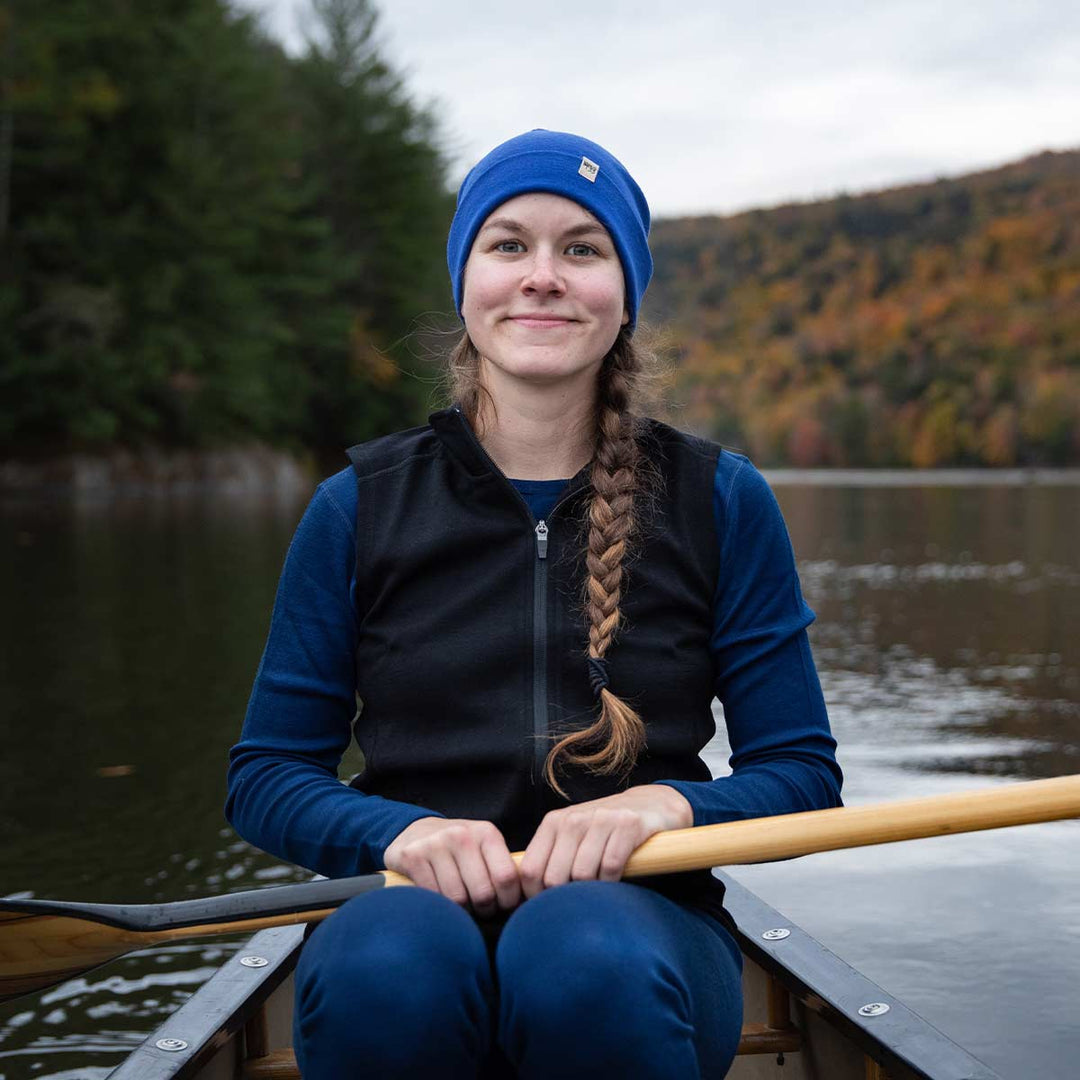 A woman sits in a canoe on open water in front of mountains and trees. The woman is wearing a lightweight hat, midweight base layer top and bottoms, and a hundred percent merino wool heavyweight full zip vest. This outer layer features a two and a half inch mock neck collar and two zippered pockets. #color_black