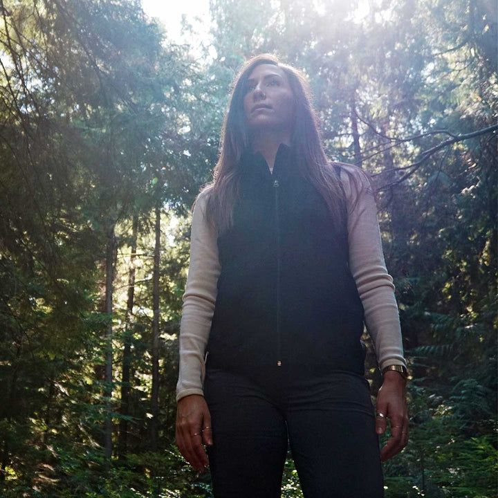 A woman in the woods models a hundred percent merino wool heavyweight full zip vest over a midweight base layer. This outer layer features a two and a half inch mock neck collar and two zippered pockets. #color_navy