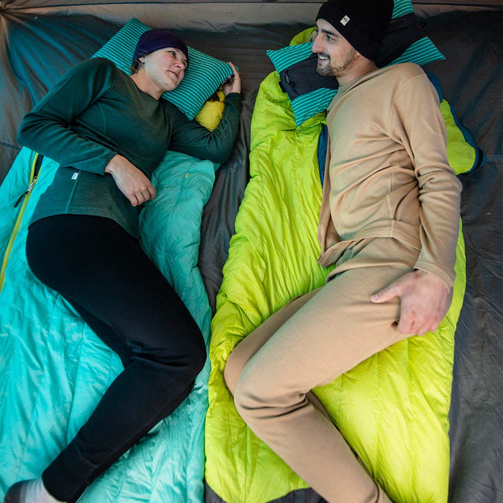 Two people lay on top of sleeping bags in a tent. Both are wearing lightweight hats and midweight base layer tops and bottoms. #color_black