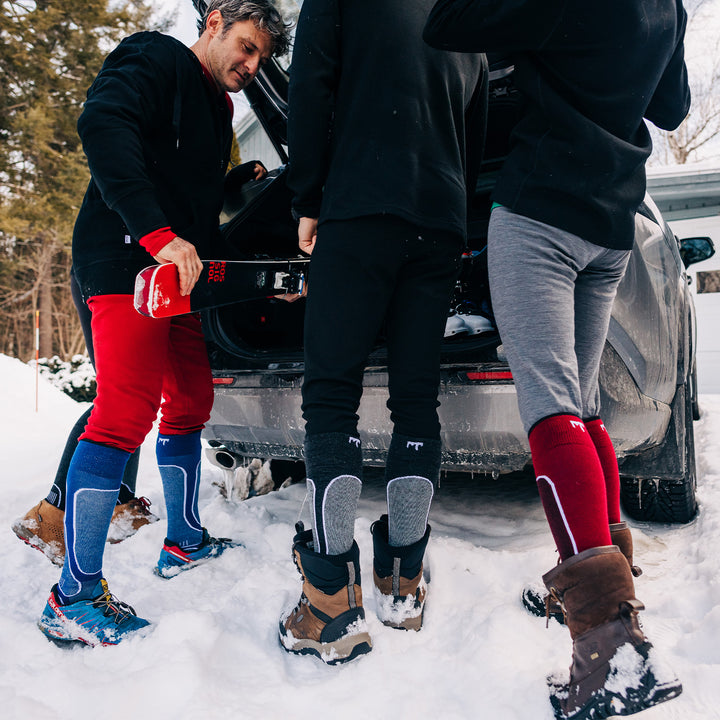 Three people stand in front of a car unloading their skis. The three people each wear a pair of one hundred percent merino wool midweight base layer bottoms, as well as ski and snowboarding socks. These bottoms feature a gusset between the legs for improved range of motion, and cuffs around the ankles for comfort and style. #color_black