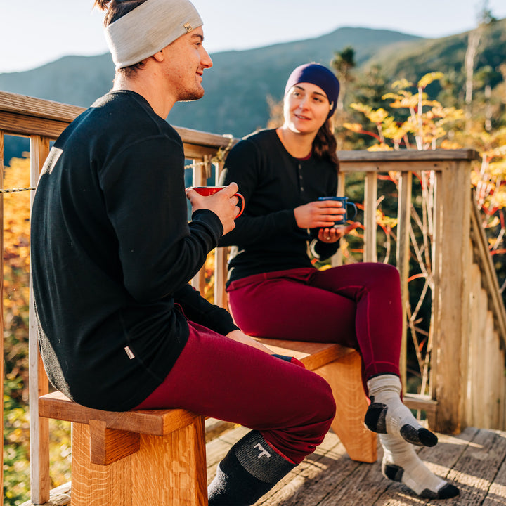 Two people sit on a porch bench with mugs in hand. Both are wearing a pair of one hundred percent merino wool midweight base layer bottoms. The women's bottoms feature a gusset between the legs for improved range of motion, and cuffs around the ankles for comfort and style. #color_burgundy