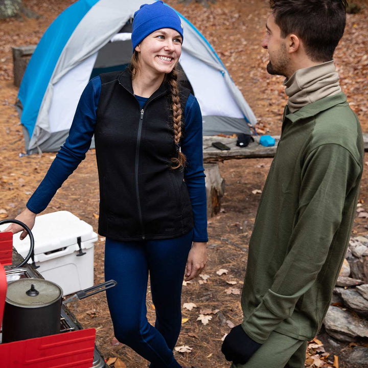 Two people chat in front of their tent and campsite. The woman wears a pair of one hundred percent merino wool midweight women's base layer bottoms. These bottoms feature a gusset between the legs for improved range of motion, and cuffs around the ankles for comfort and style. #color_navy