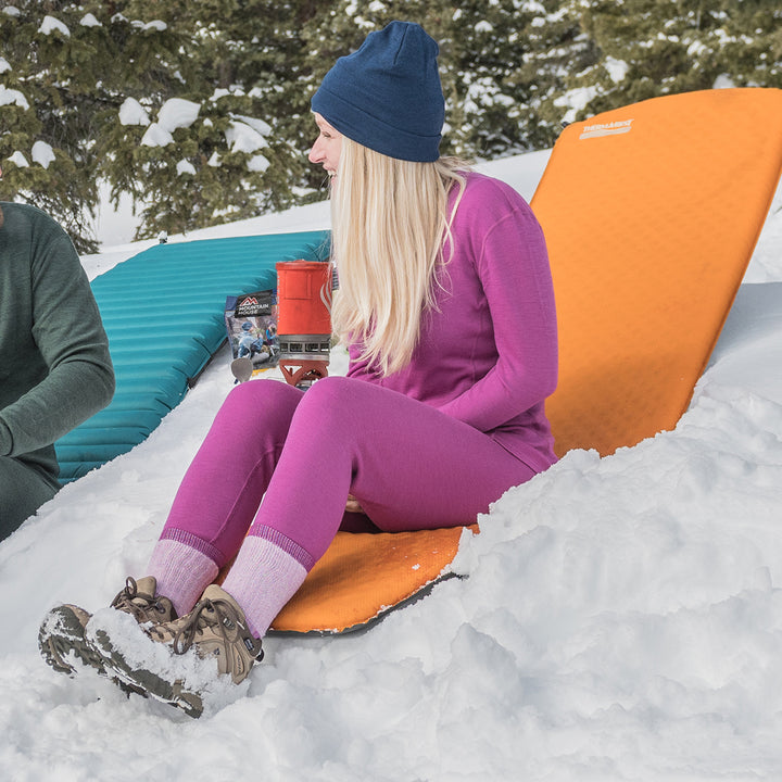 A woman sits on a chair in the snow wearing a full base layer set. She models a hat, a base layer crew top, a pair of one hundred percent merino wool midweight women's base layer bottoms, and a pair of heavyweight socks. The bottoms feature a gusset between the legs for improved range of motion, and cuffs around the ankles for comfort and style. #color_radiant-violet