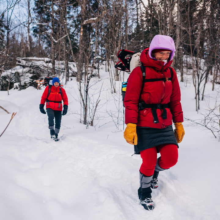 Two people hike a snowy trail in parkas and gloves. The woman in front is wearing a pair of one hundred percent merino wool midweight women's base layer bottoms. These bottoms feature a gusset between the legs for improved range of motion, and cuffs around the ankles for comfort and style. #color_true-red
