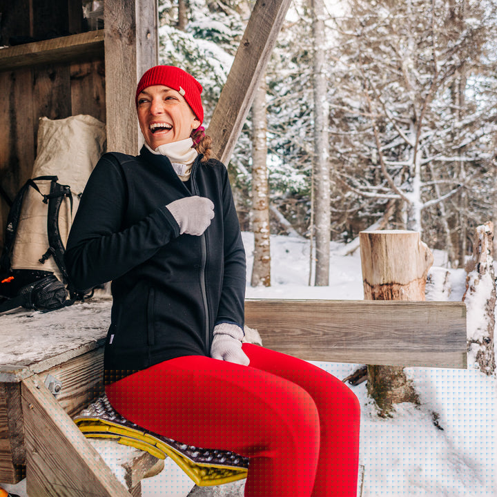 A woman sits on the front step of the camp lodge to adjust her heavyweight full zip sweater. The woman is modeling a pair of one hundred percent merino wool midweight women's base layer bottoms. These bottoms feature a gusset between the legs for improved range of motion, and cuffs around the ankles for comfort and style. #color_true-red